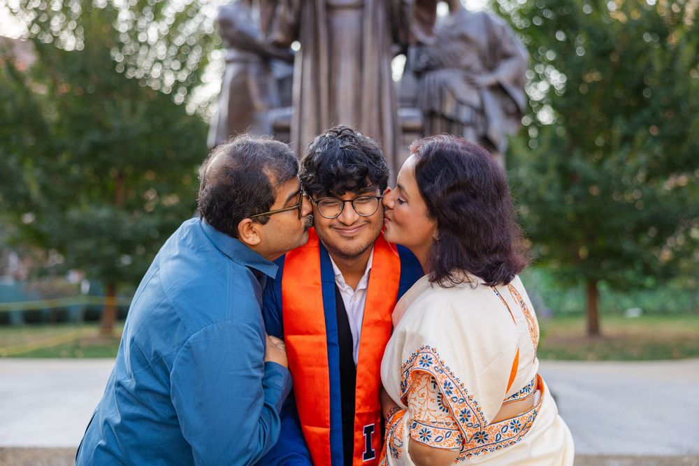 Graduating student wearing graduation cap and gown at Alma Mater getting a kiss on the cheek from his parents