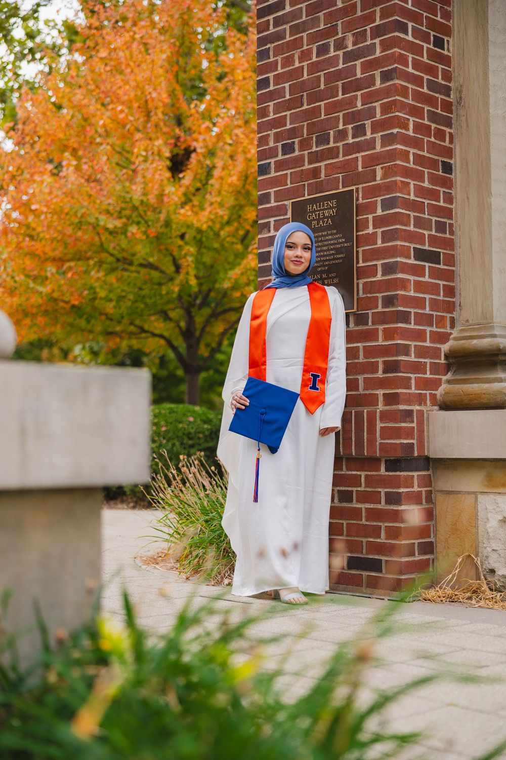 Student with cap and stole leaning on brick wall at Hallene Gateway in the fall