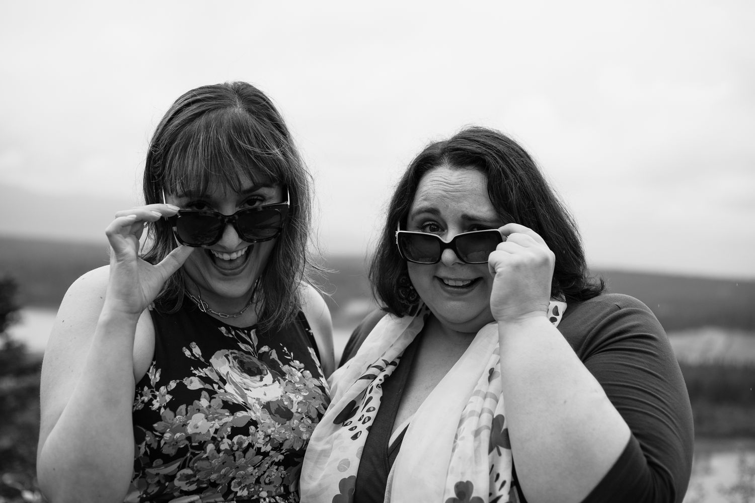 Black and white photo of two women laughing while wearing sunglasses outdoors.