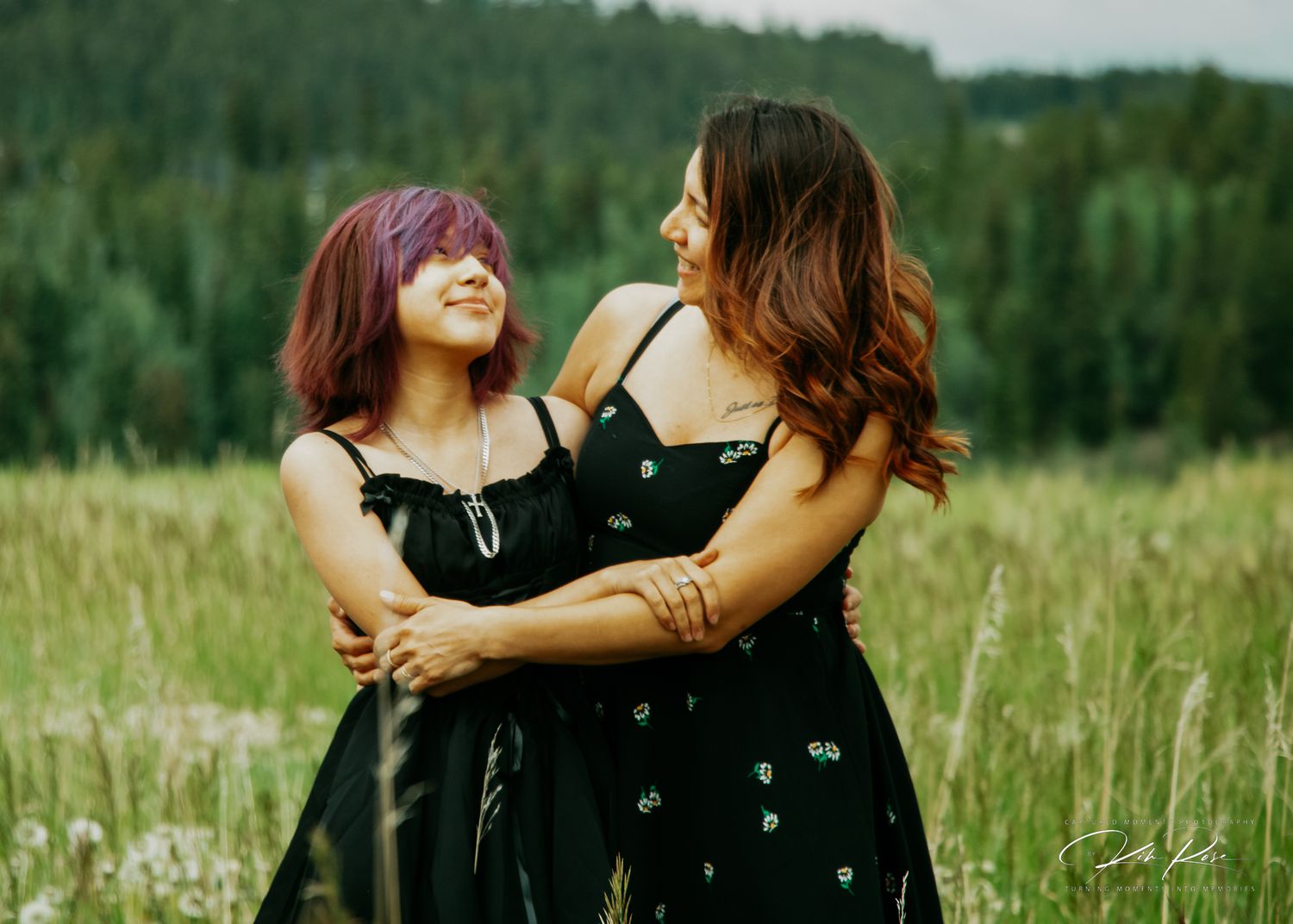 Two young women in black dresses embracing and laughing in a meadow.