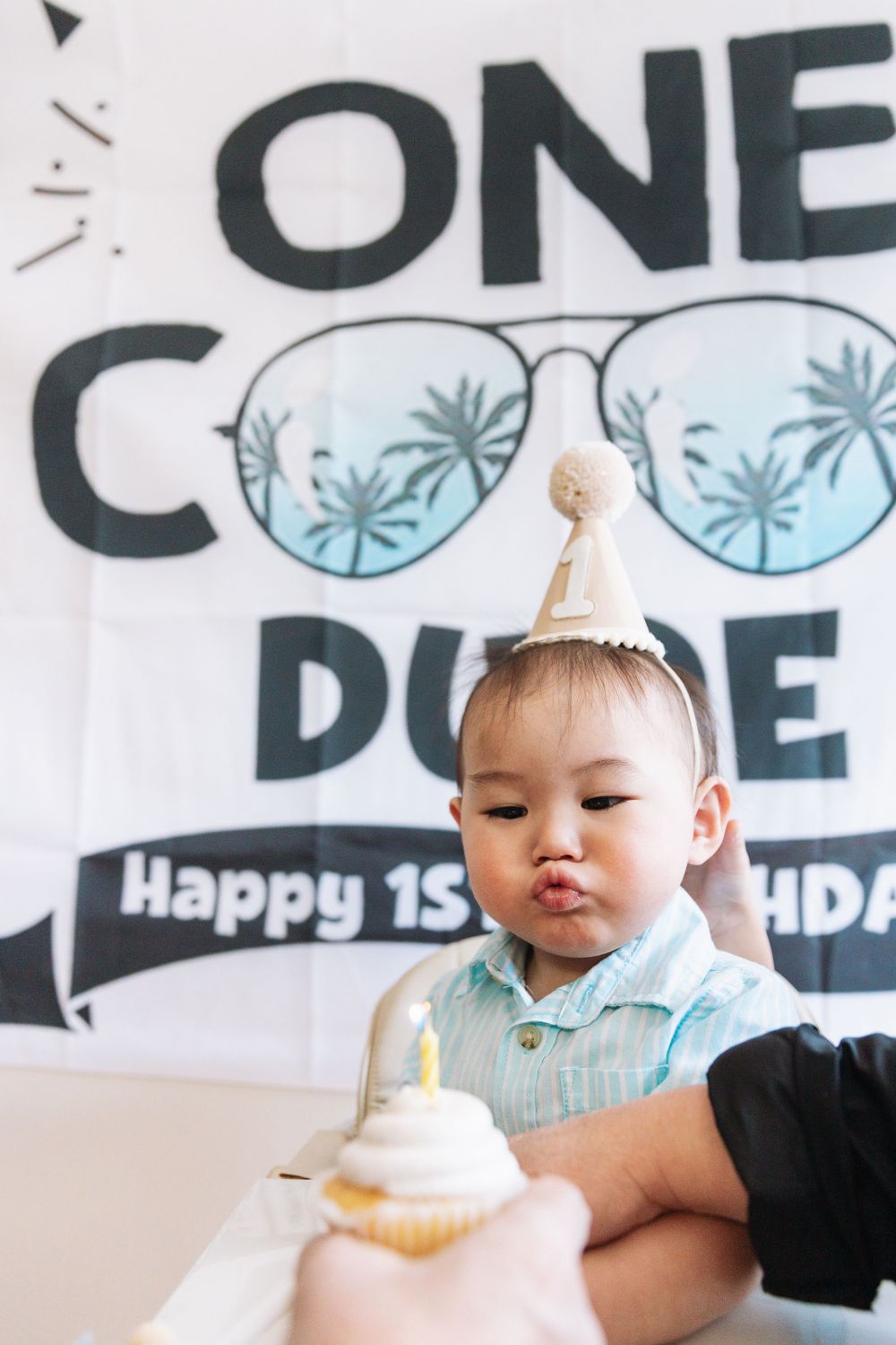 A baby celebrates their first birthday with cake against a decorated backdrop with sunglasses design.