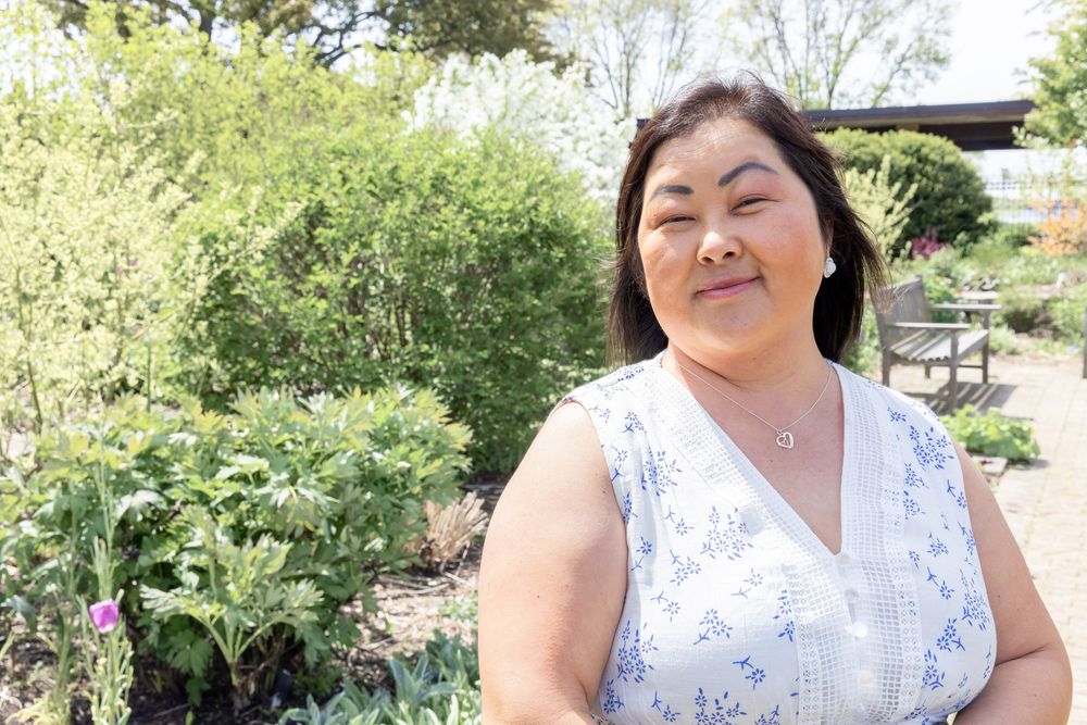 A woman in a floral dress poses in a sunny garden setting with blooming flowers.
