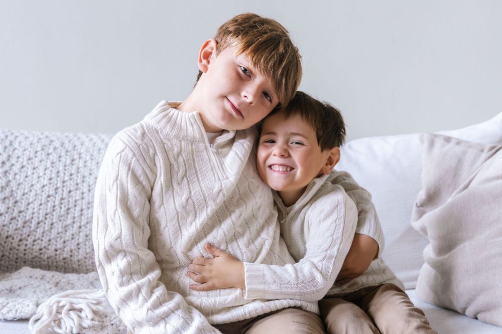 Two children in white sweaters cuddling on white bedding.