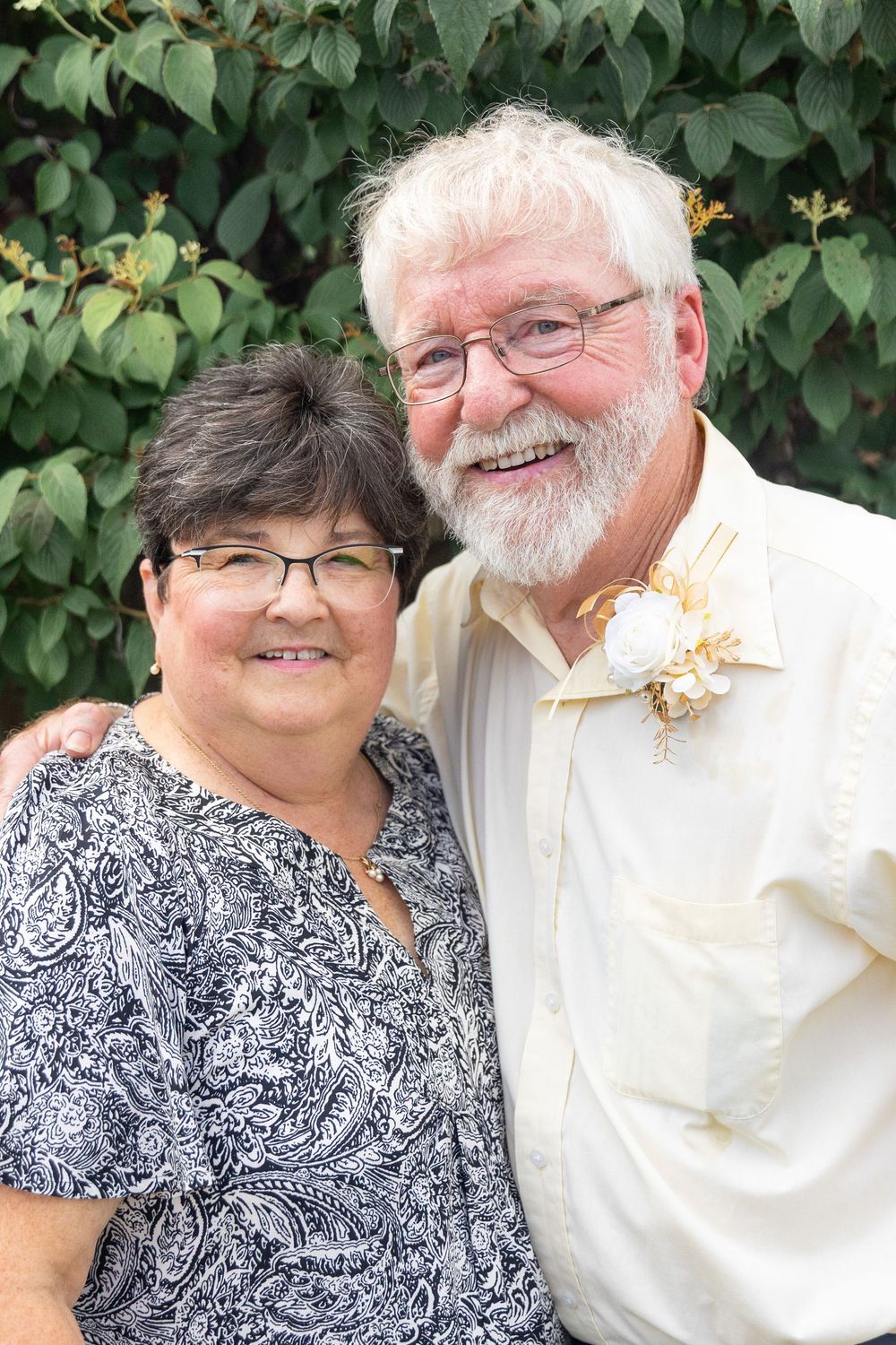 Smiling elderly couple posing outdoors, man with white beard wearing boutonniere, woman in paisley blouse, green foliage background.