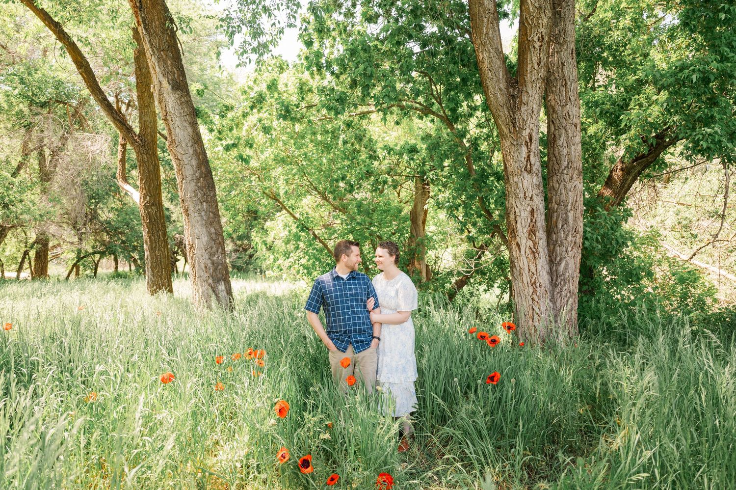 A couple standing among wildflowers and tall trees in a sunlit summer meadow.
