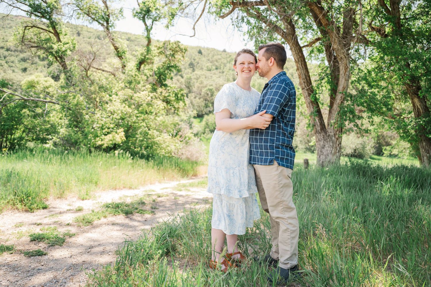 Romantic couple embrace on a sunlit nature trail surrounded by lush green trees and foliage.