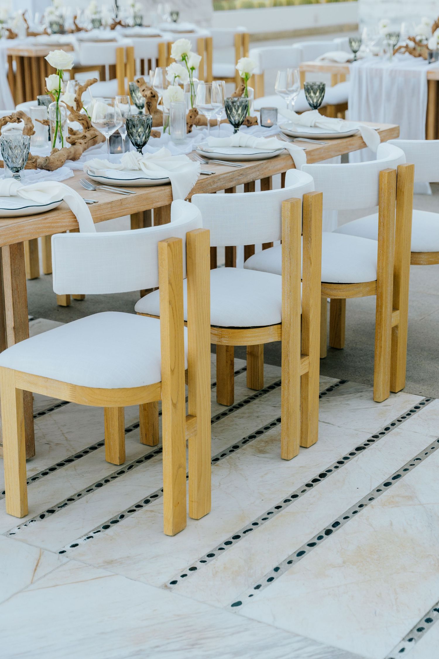 Elegant outdoor wedding reception tables with white upholstered wooden chairs, floral centerpieces, and glassware on a tiled patio.