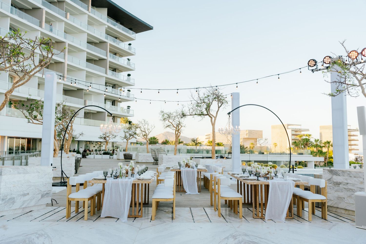 Elegant outdoor wedding reception with wooden tables, white linens and string lights on a modern resort terrace overlooking the coast.