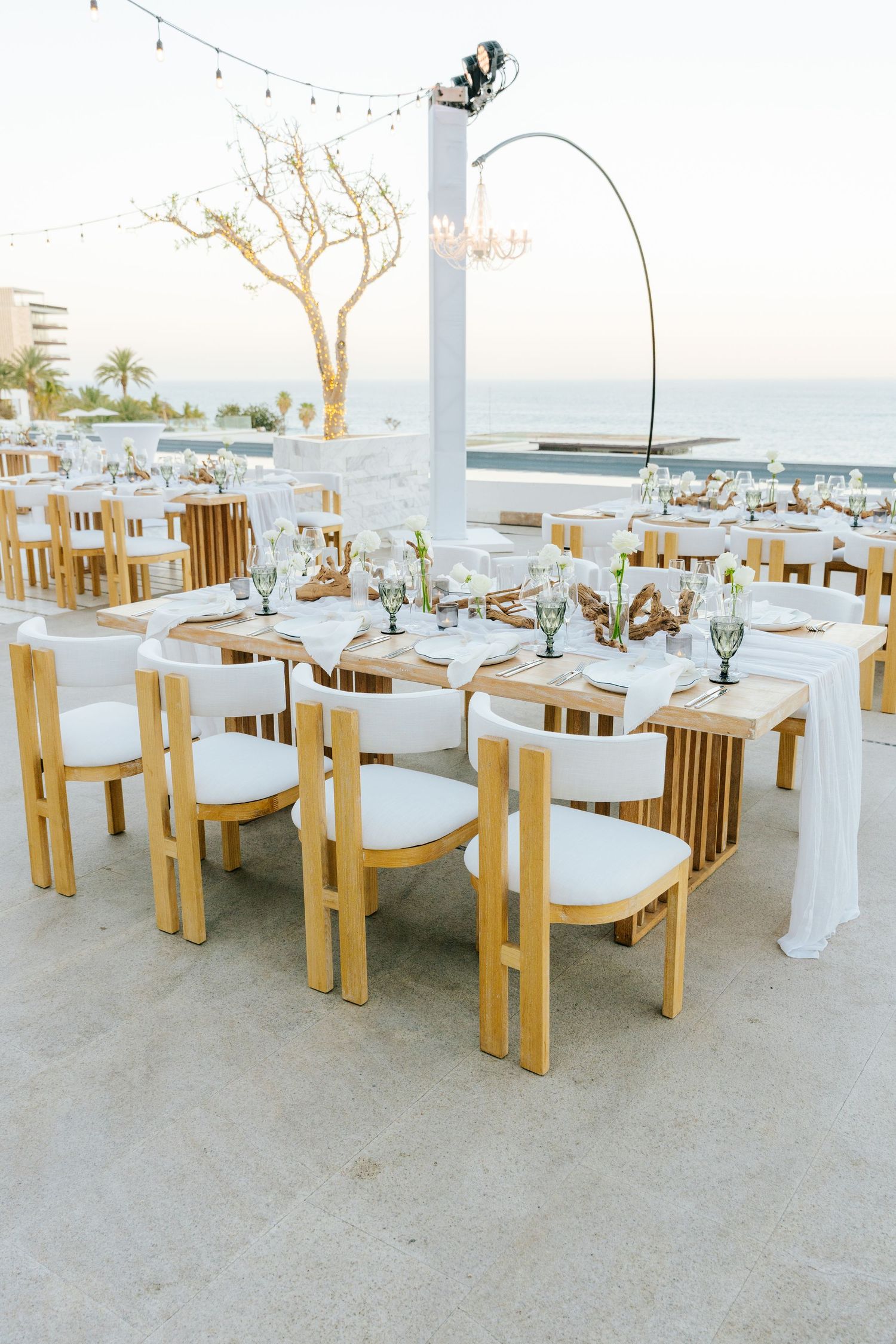 Elegant oceanfront wedding reception with wooden tables, white cushioned chairs, glassware, and chandeliers on a seaside terrace at sunset.