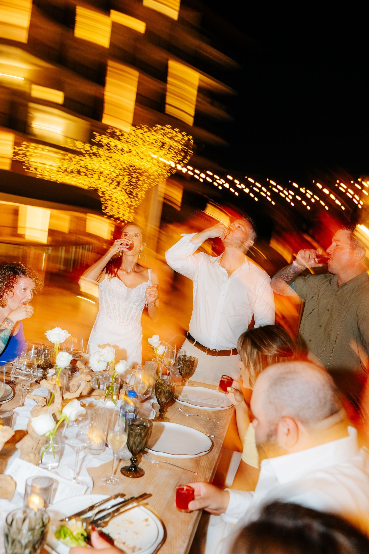Wedding guests raise drinks in a lively toast around a candlelit outdoor dinner table at night, with glowing string lights behind.