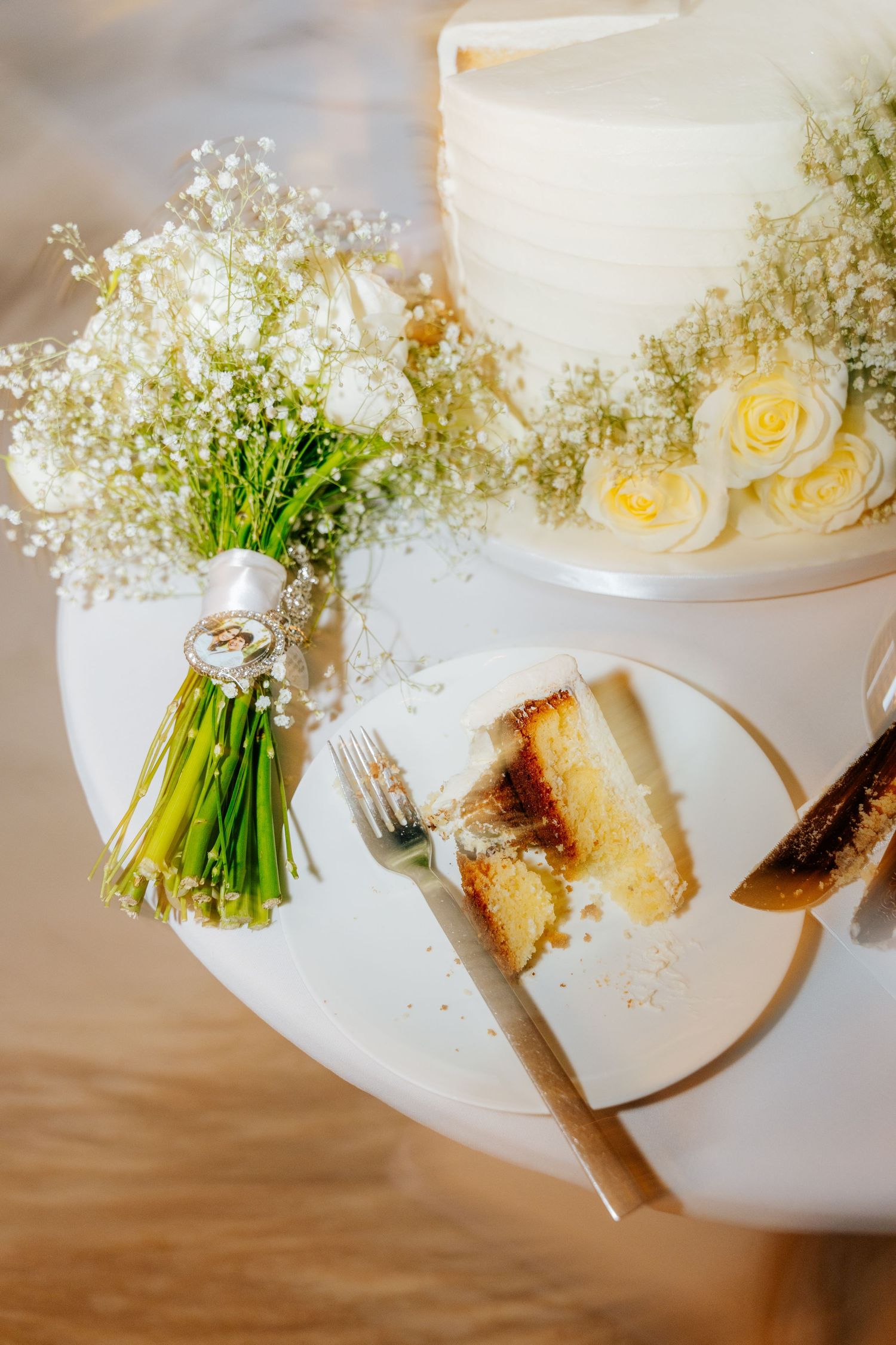 Wedding bouquet of baby's breath with rings on the ribbon beside a sliced white cake with yellow roses and a slice on a plate.