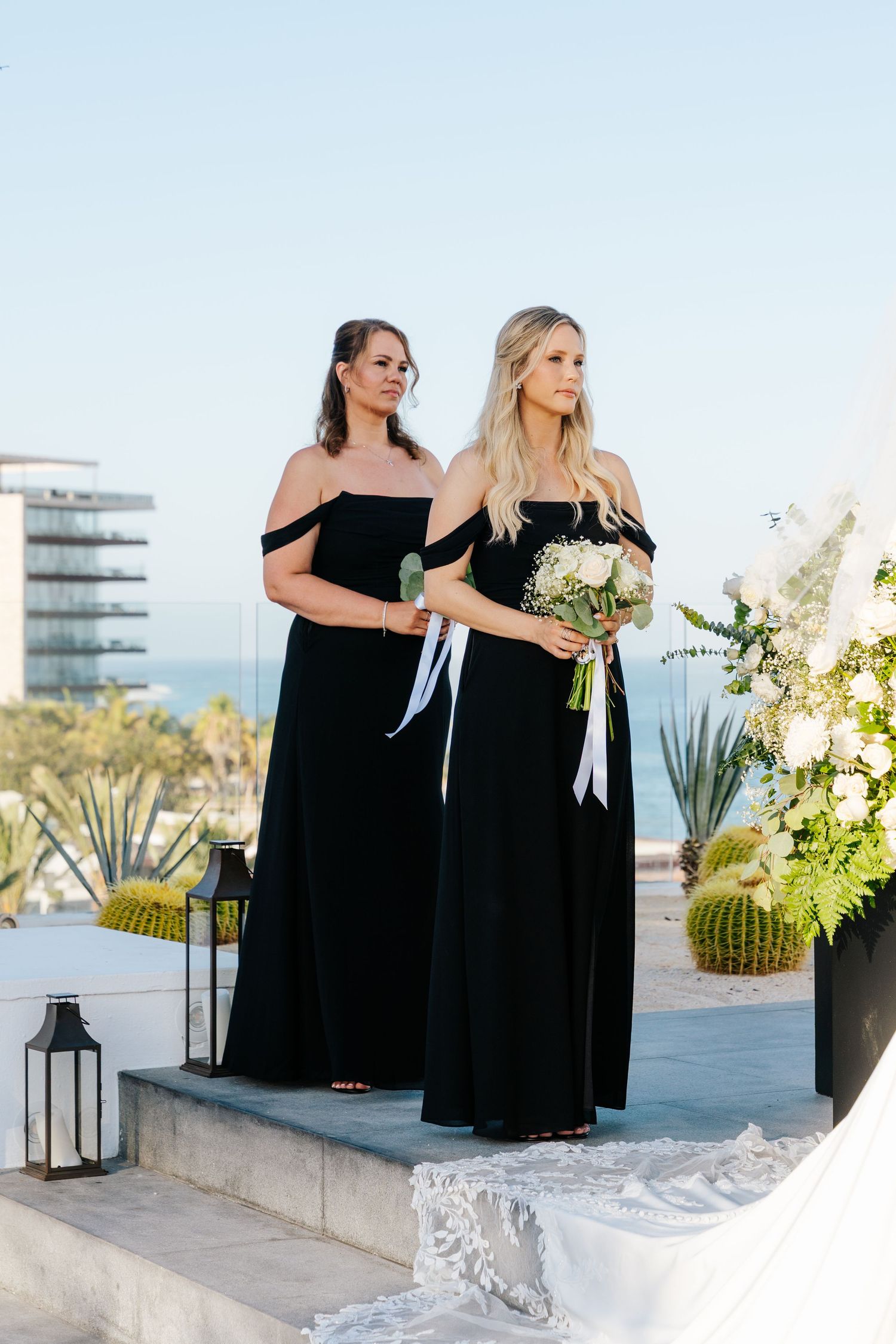 Two bridesmaids in black off-the-shoulder gowns hold white bouquets on a rooftop wedding ceremony space with ocean views and lace veil.