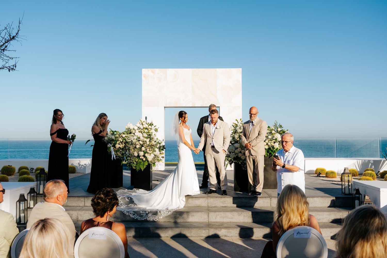 Bride and groom exchange vows during an outdoor oceanfront wedding ceremony with seated guests, bridesmaids, and floral arrangements.