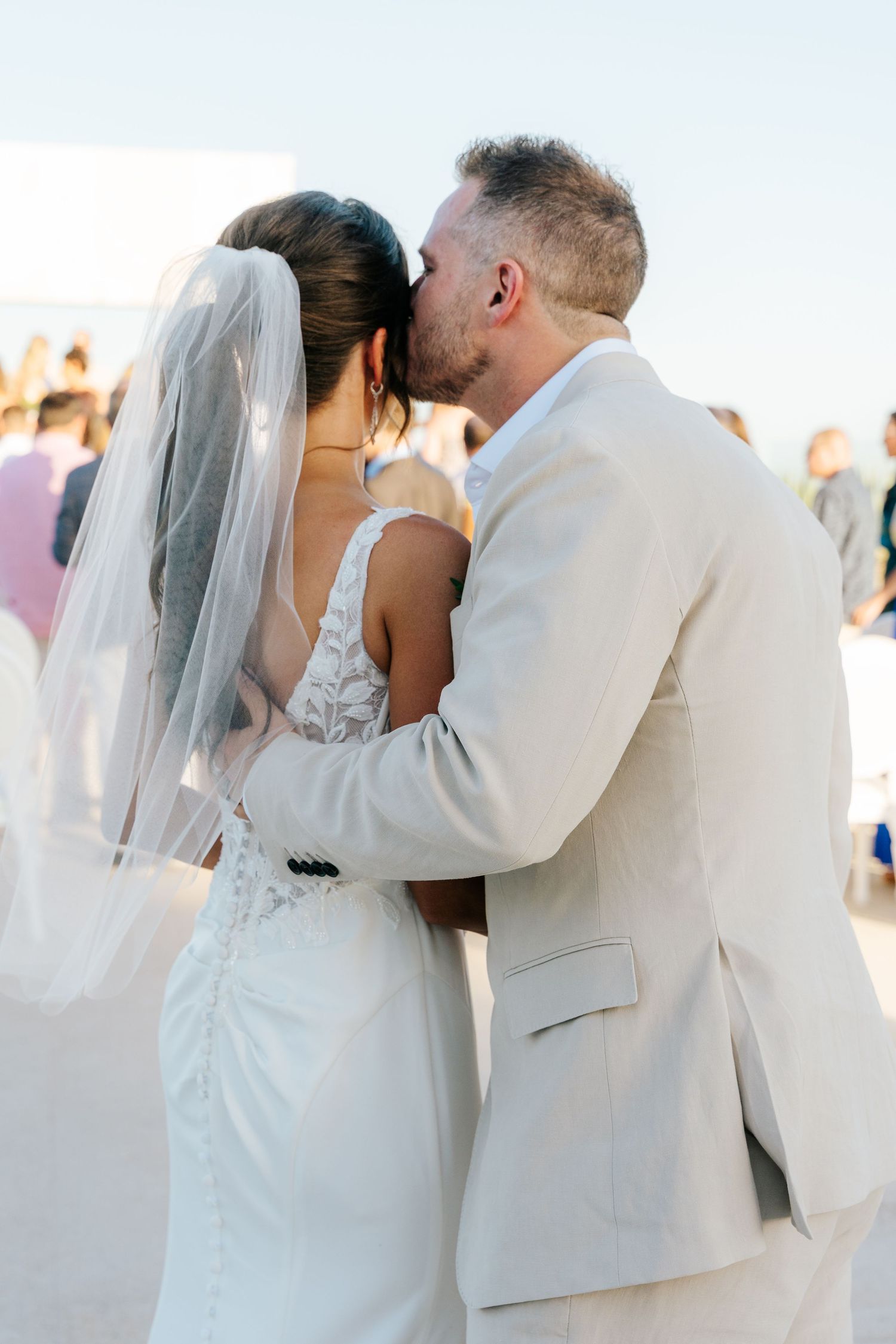A groom in a light beige suit kisses the forehead of his bride in a lace gown and long veil at an outdoor beach wedding.