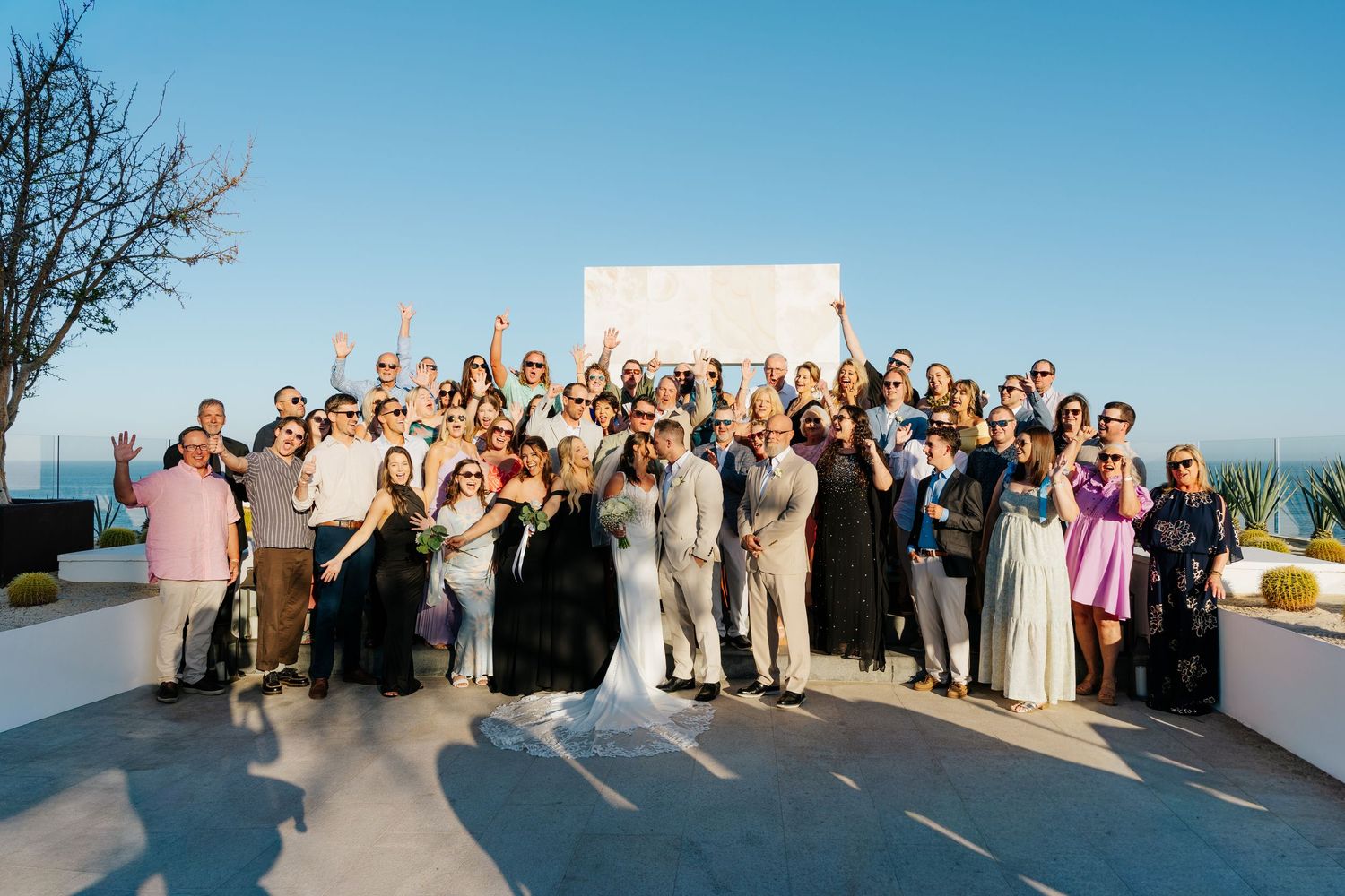 Large wedding party and guests cheer together for a group photo on a sunny seaside terrace with the bride and groom at the center.