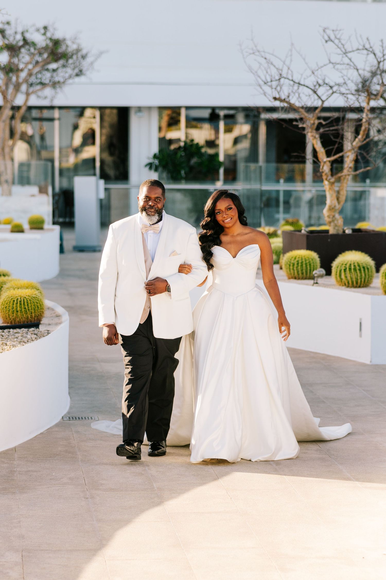 A bride and groom in white wedding attire pose on a modern rooftop terrace surrounded by barrel cacti.