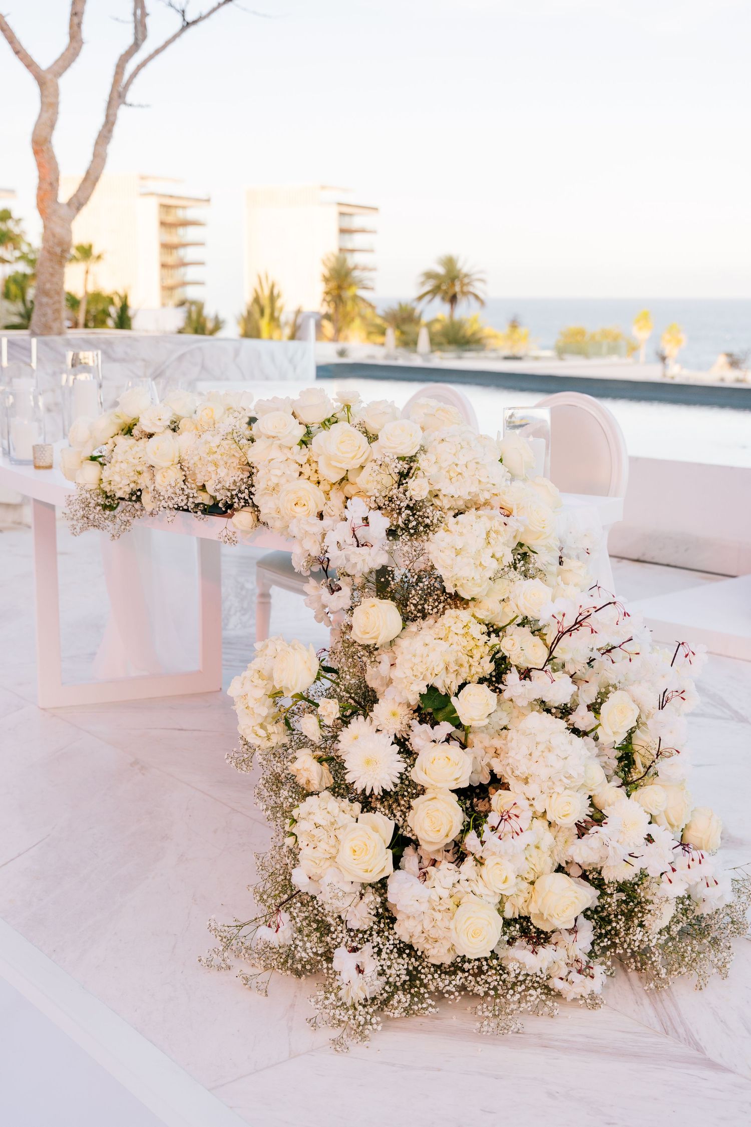 Elegant outdoor wedding table adorned with lush white roses, hydrangeas, and baby's breath arrangements against ocean backdrop.