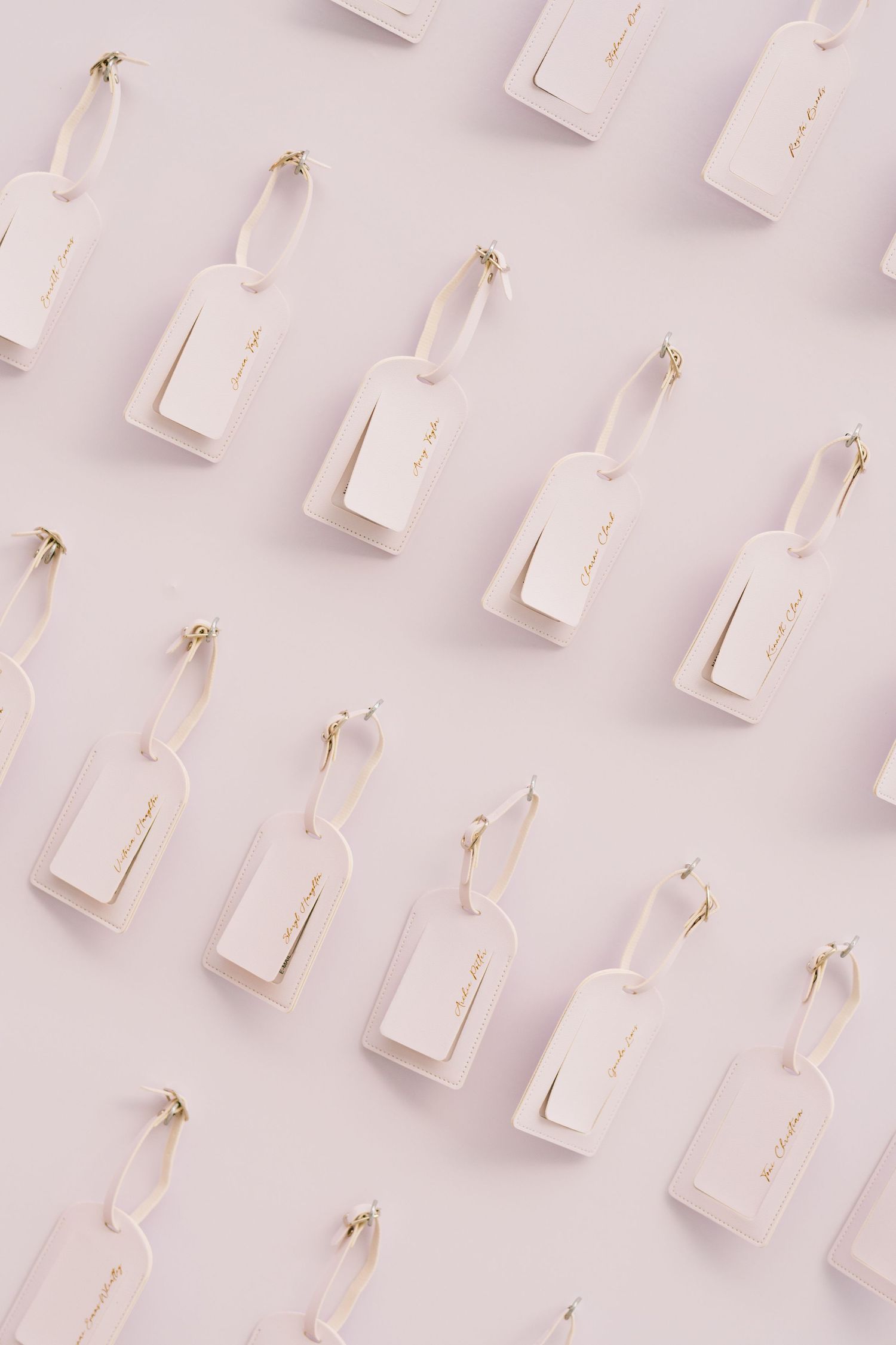 Elegant white luggage tags with gold script and ribbon loops arranged in a diagonal pattern on a soft pink background.