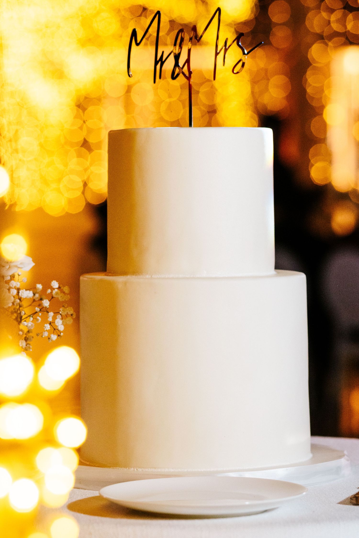 Elegant two-tier white wedding cake with Mrs & Mrs topper, set against warm golden bokeh lights.