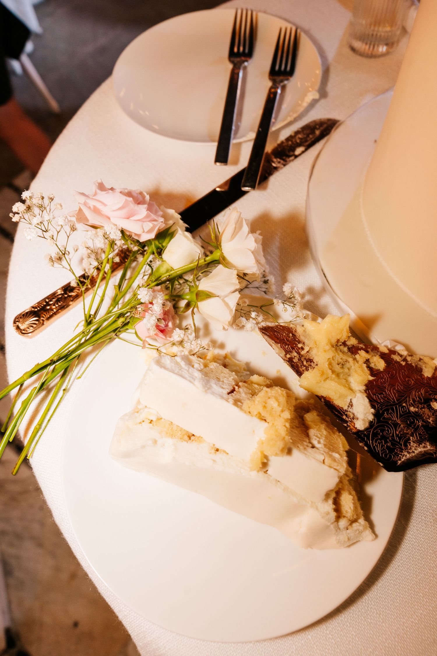 Elegant wedding cake slice on white plate with pink roses and gold cutlery at a reception dinner.