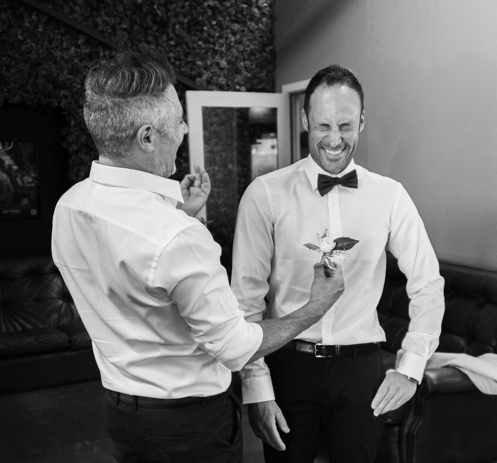 Two people in formal attire share a laugh during wedding preparations.