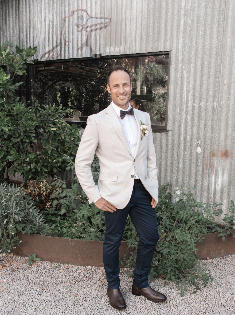 Person in cream tuxedo jacket and bow tie poses against industrial wall with greenery.