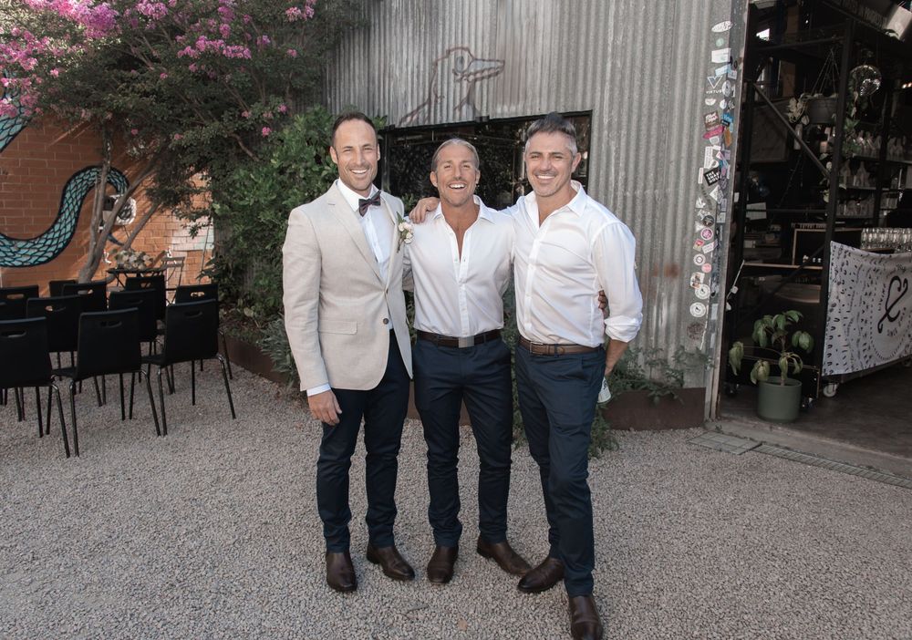 Three people in light-colored business attire posing together at an outdoor venue with tables and decorative flowers.
