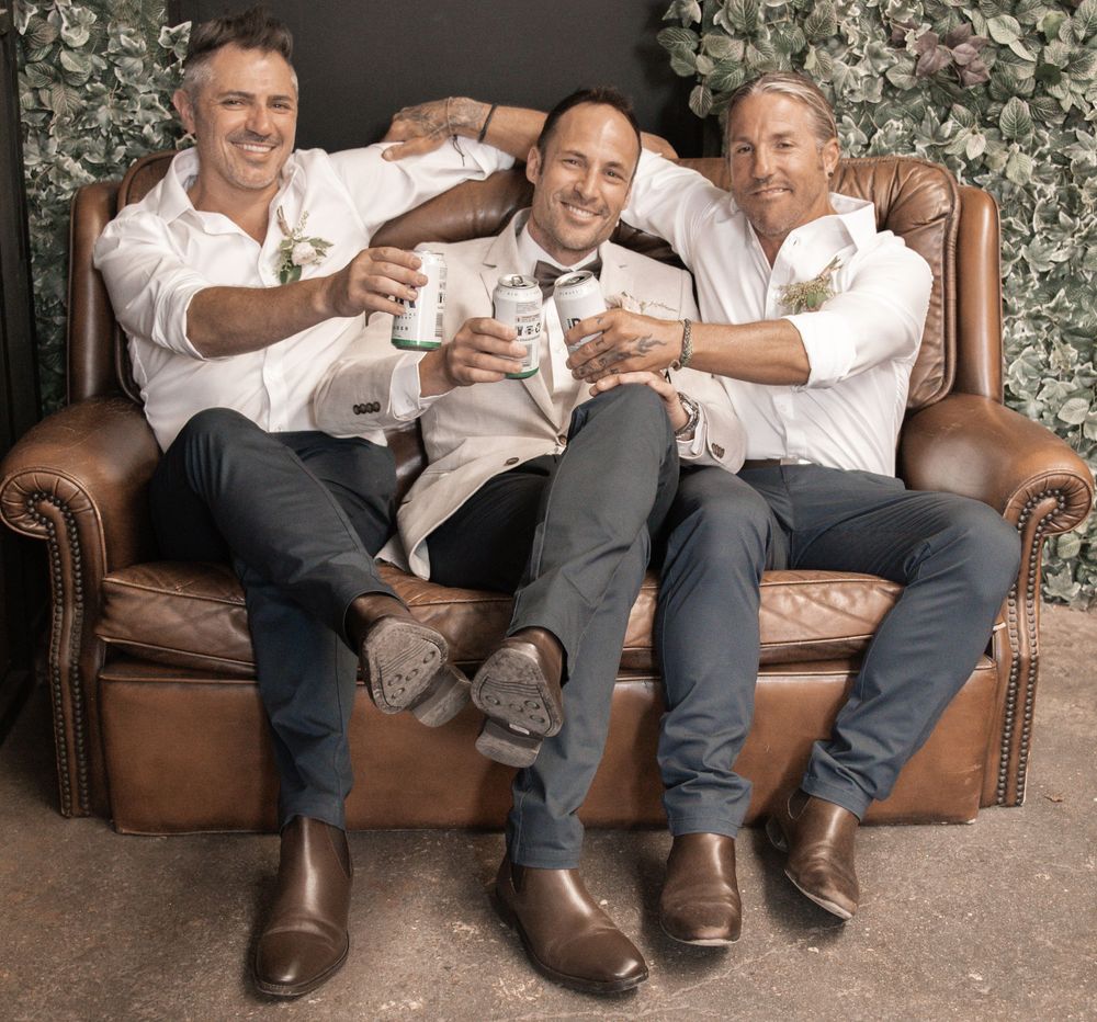 Three people toasting with drinks while sitting on a brown leather couch against a floral backdrop.