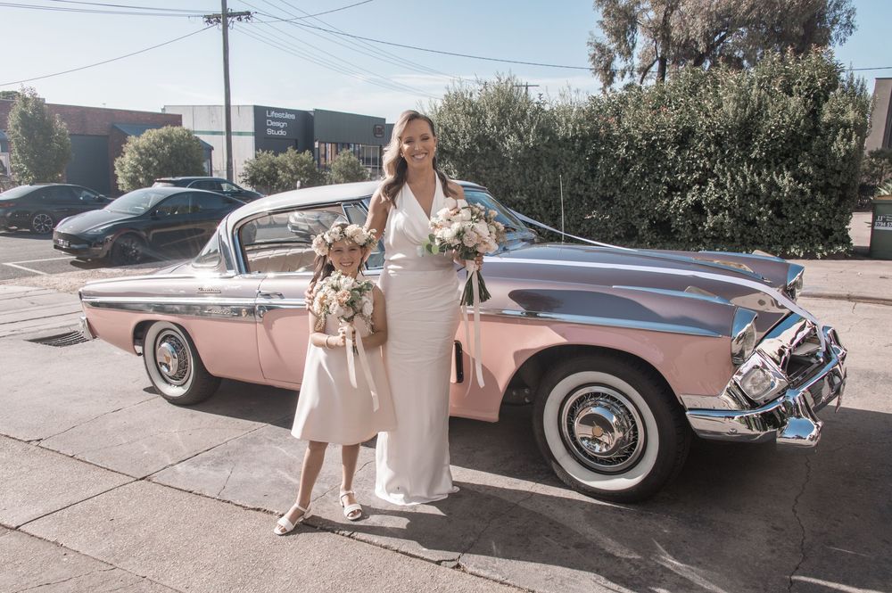 Two people in elegant white attire pose beside a vintage pink car on a sunny street.