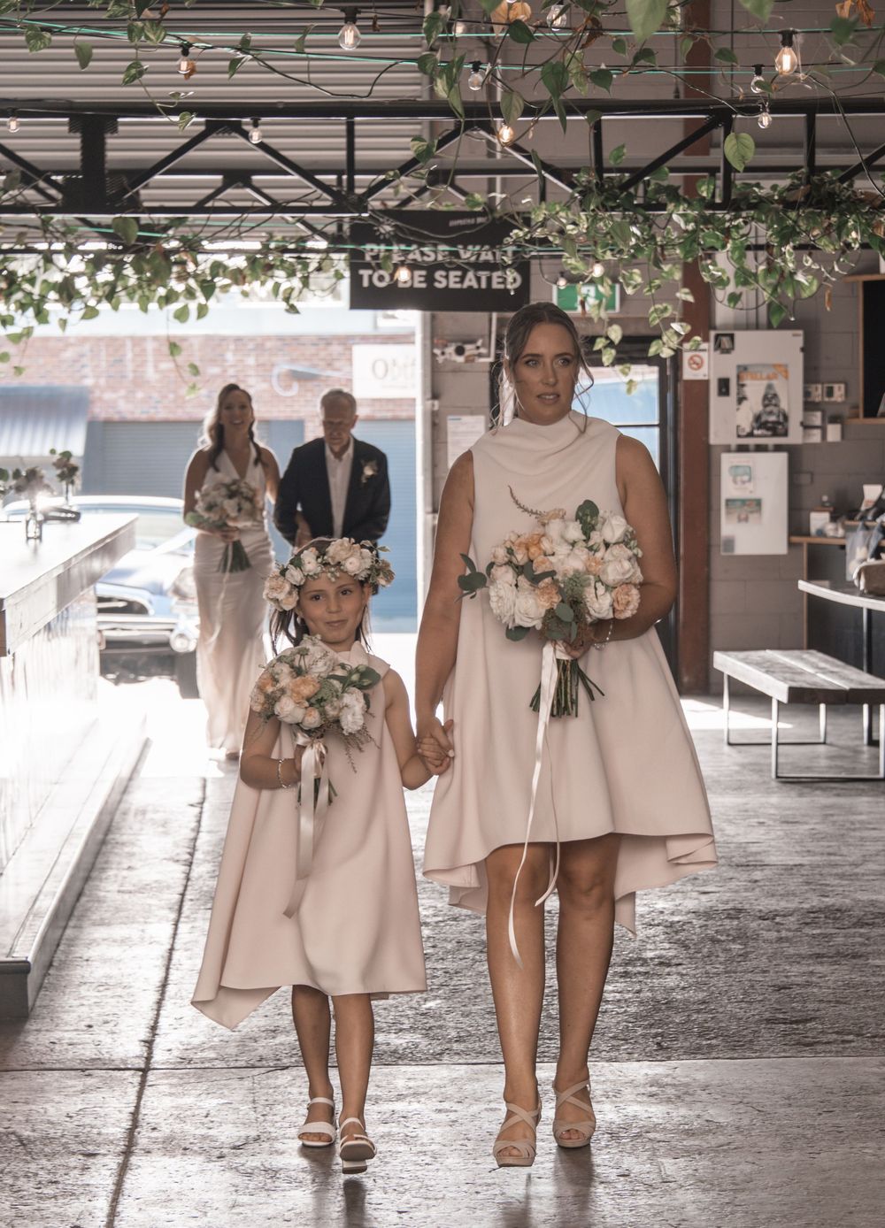 Two people in blush pink dresses walk together holding bouquets under industrial ceiling.