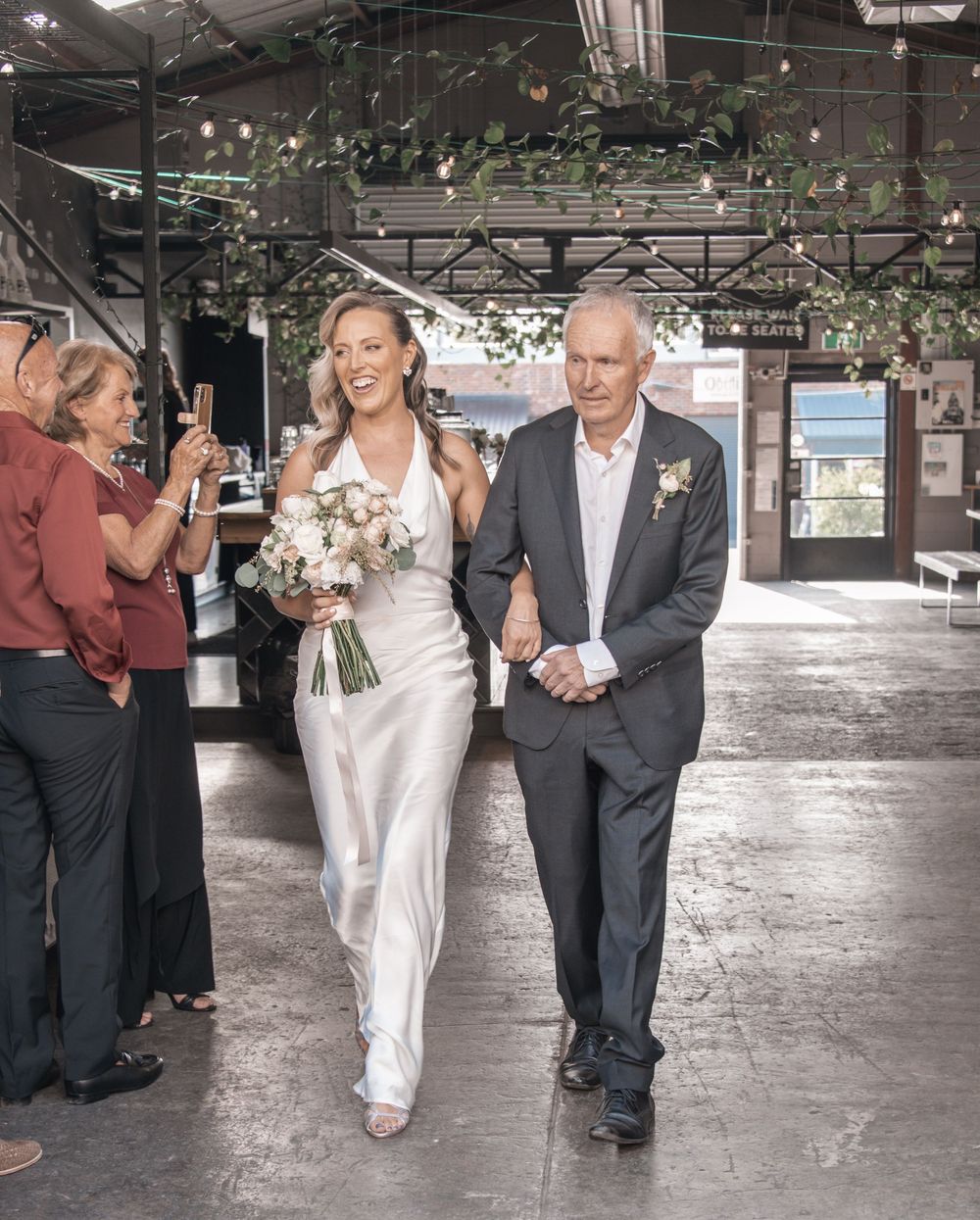 A wedding procession with the bride in white and her escort walking down an industrial-style aisle with hanging greenery.