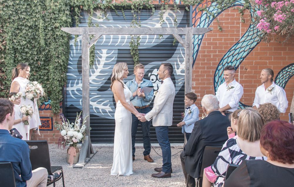 Intimate outdoor wedding ceremony under a white pergola with brick backdrop and floral arrangements.