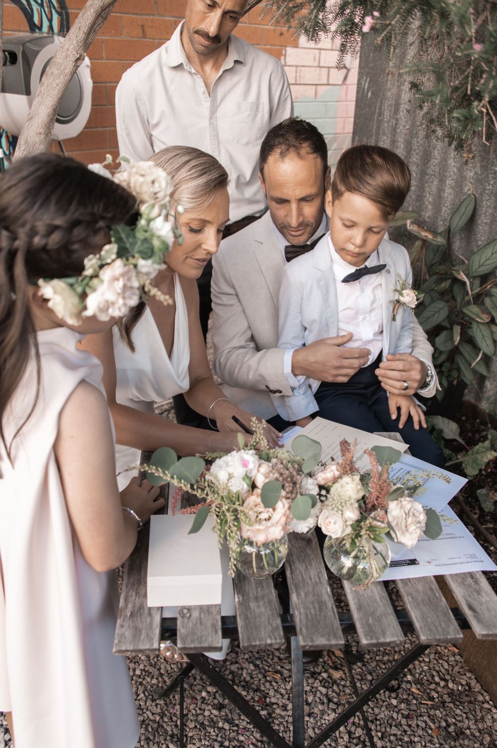Wedding party gathers around table examining floral arrangements and documents outdoors.