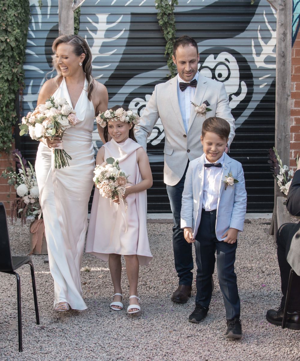 A wedding party including a bride in white, flower girl in pink, and two participants in formal attire near a mural.