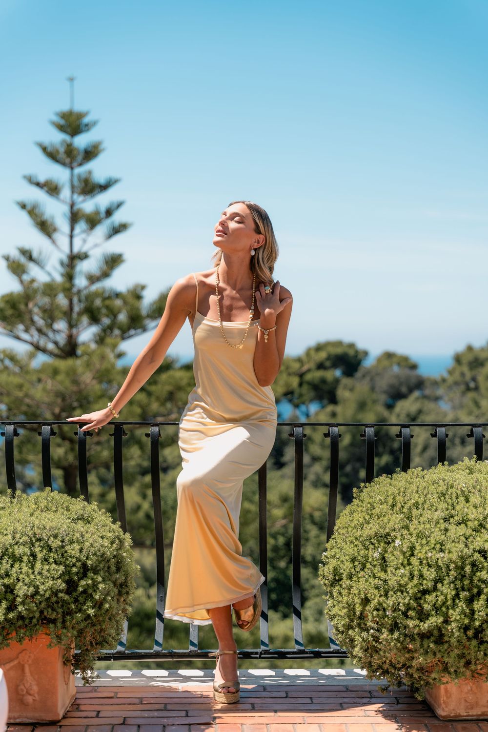 Woman in yellow dress poses gracefully against railing on sunny terrace with green plants and blue sky in background.