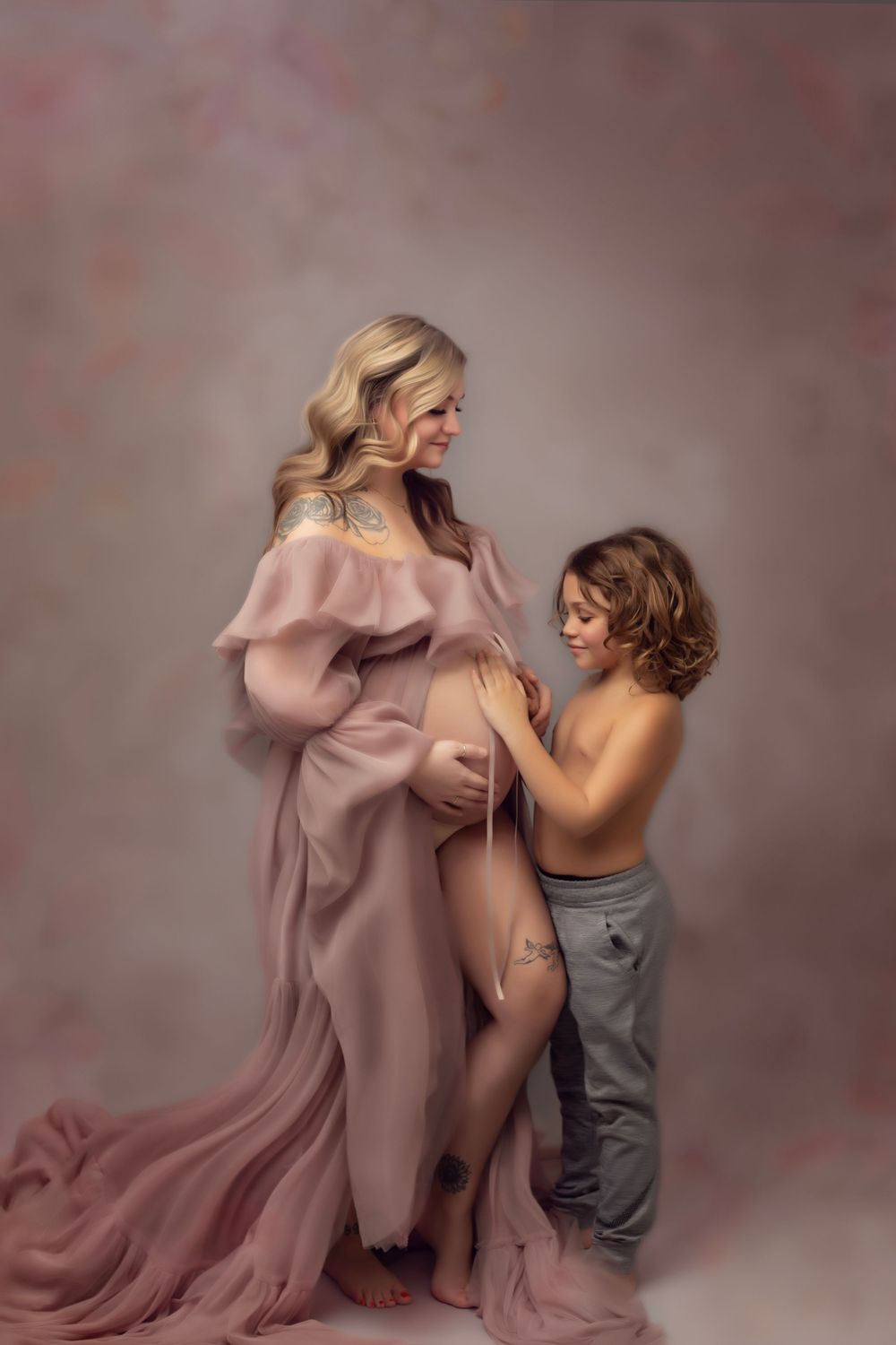A fine art maternity portrait showing a tender moment between a pregnant mother in a soft pink dress and her young son admiring Mama's belly