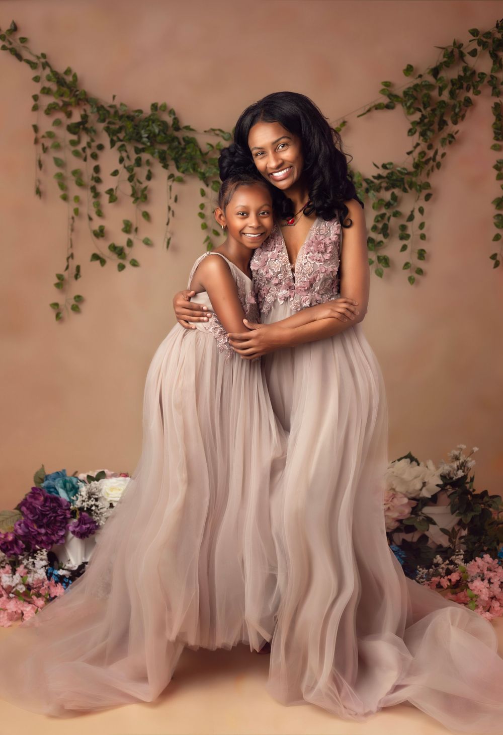 Two figures in flowing tulle dresses pose together against a neutral backdrop with floral accents.