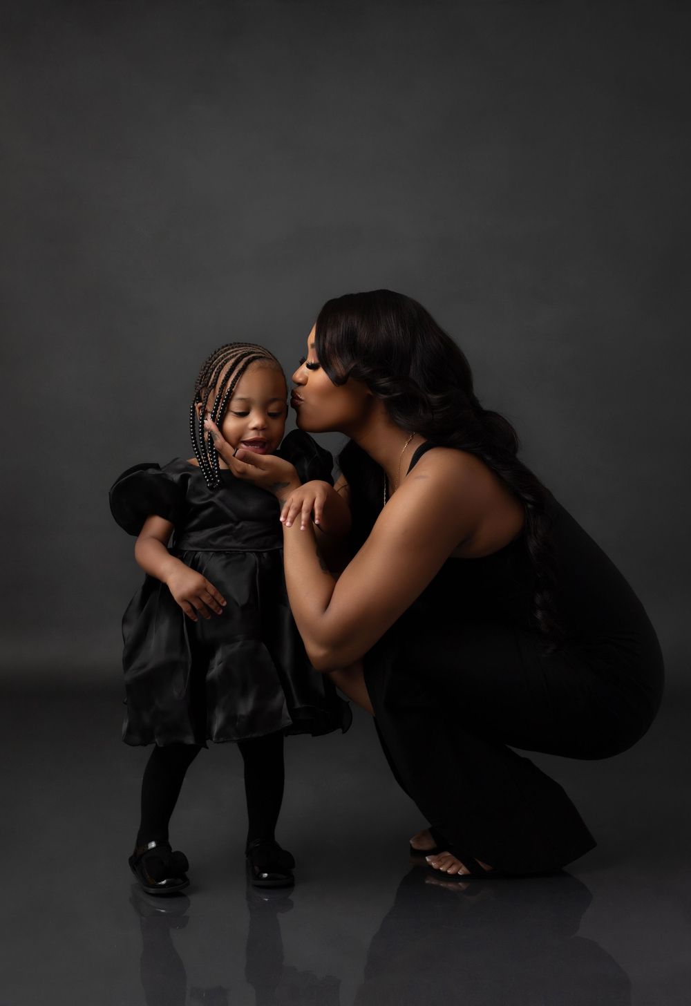 A tender moment captured in a dark studio as a woman in black dress kneels to kiss her sweet baby girl in matching dresses.