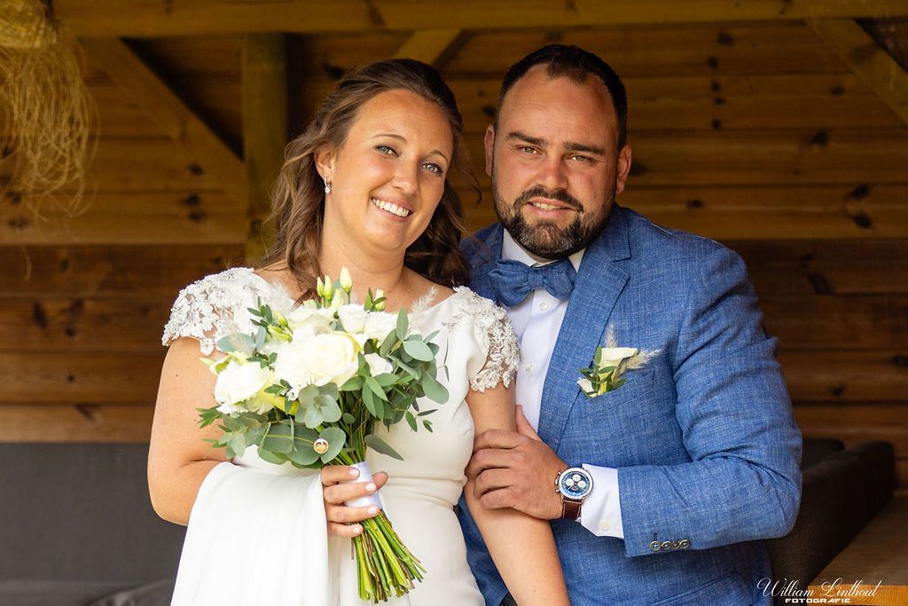 A newlywed couple poses with a white floral bouquet under a wooden shelter.