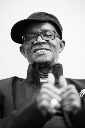 A black and white portrait of someone wearing glasses and a cap smiling warmly while looking down. Beres Hammond