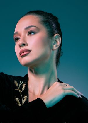 Portrait photo with dramatic blue lighting showing upturned face wearing a black outfit with gold leaf embroidery detail.