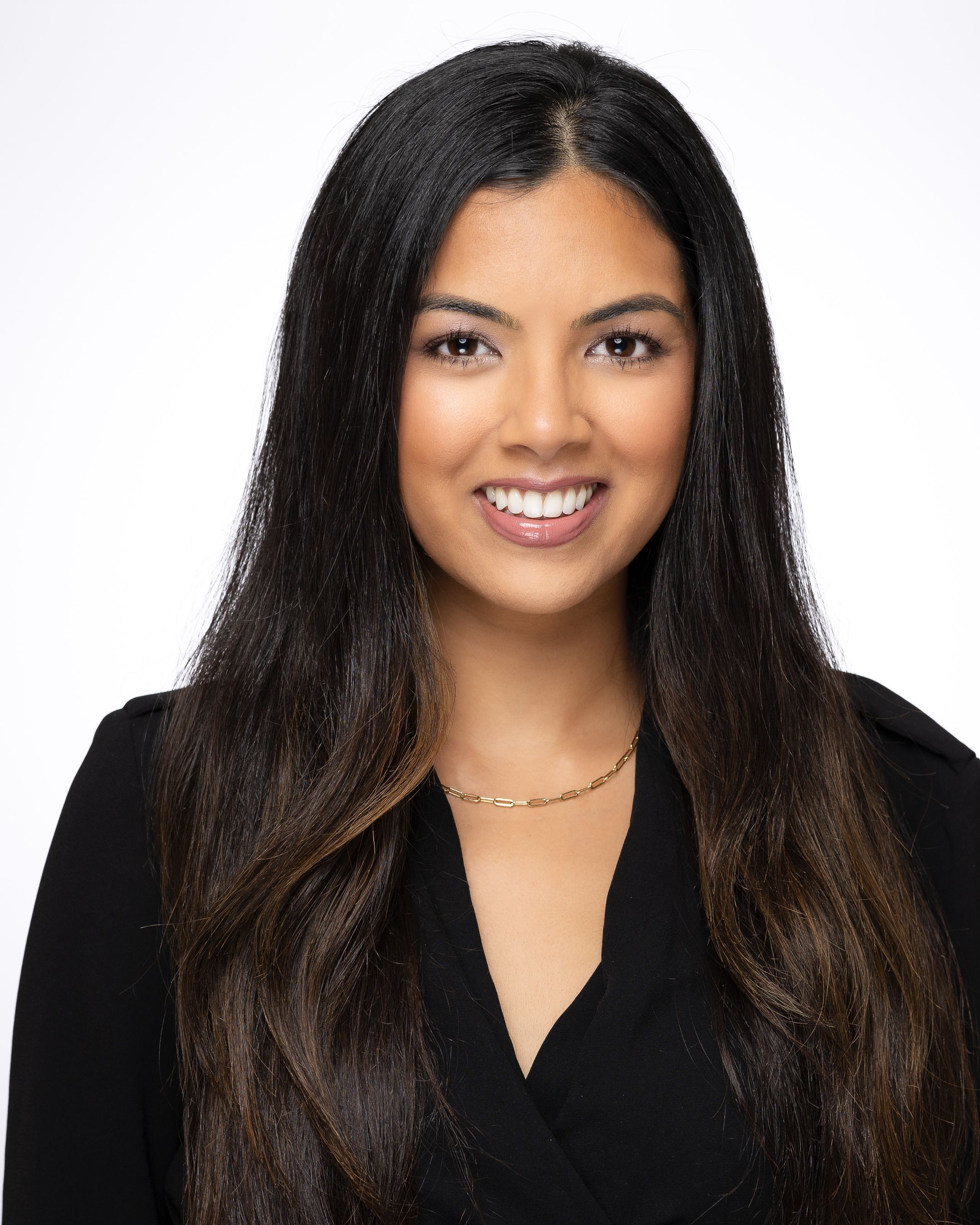 Professional headshot of a person in a black top with long dark hair, necklace and bright smile against light background.