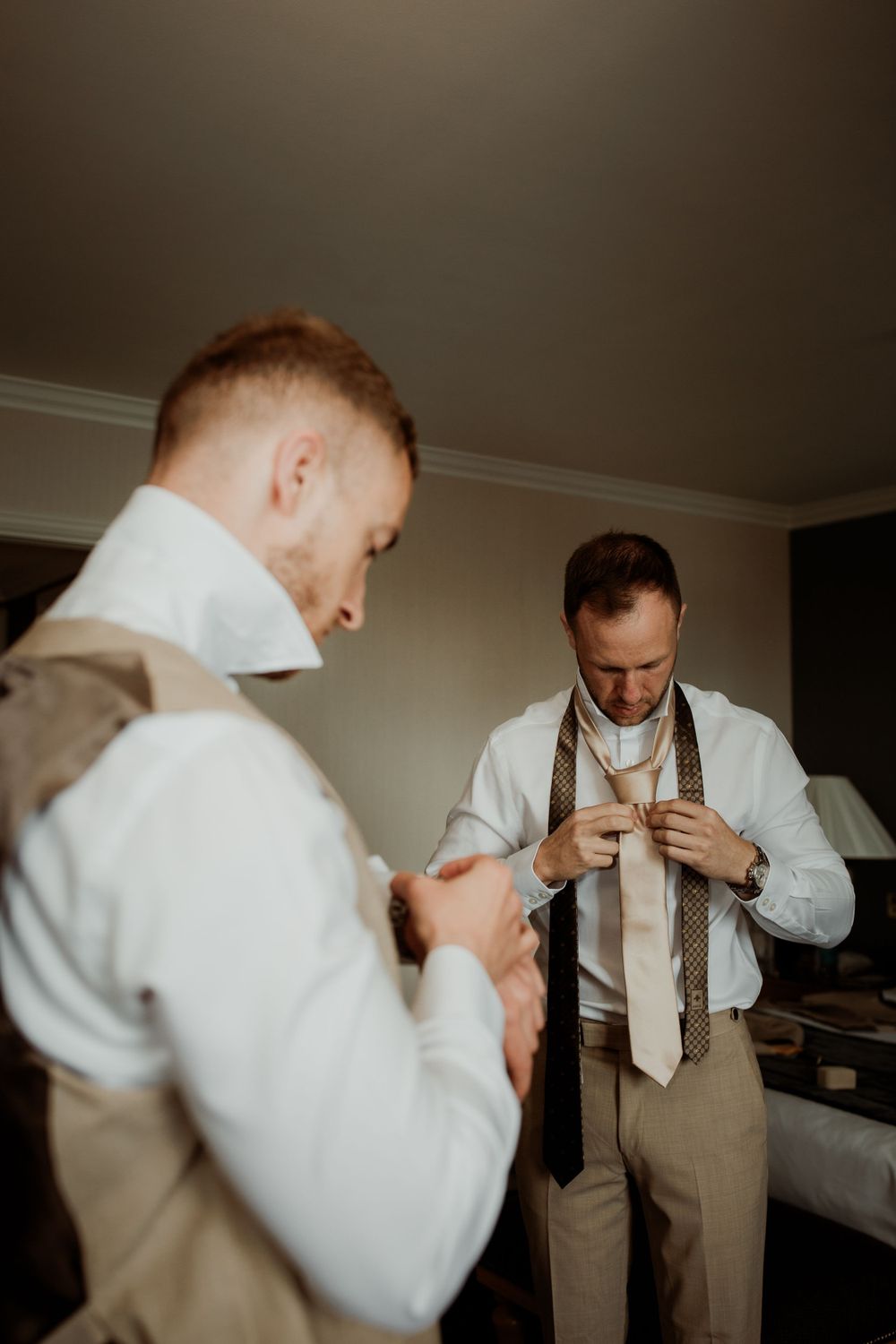 Two people getting dressed in formal attire with white shirts and tan suspenders preparing for an event.