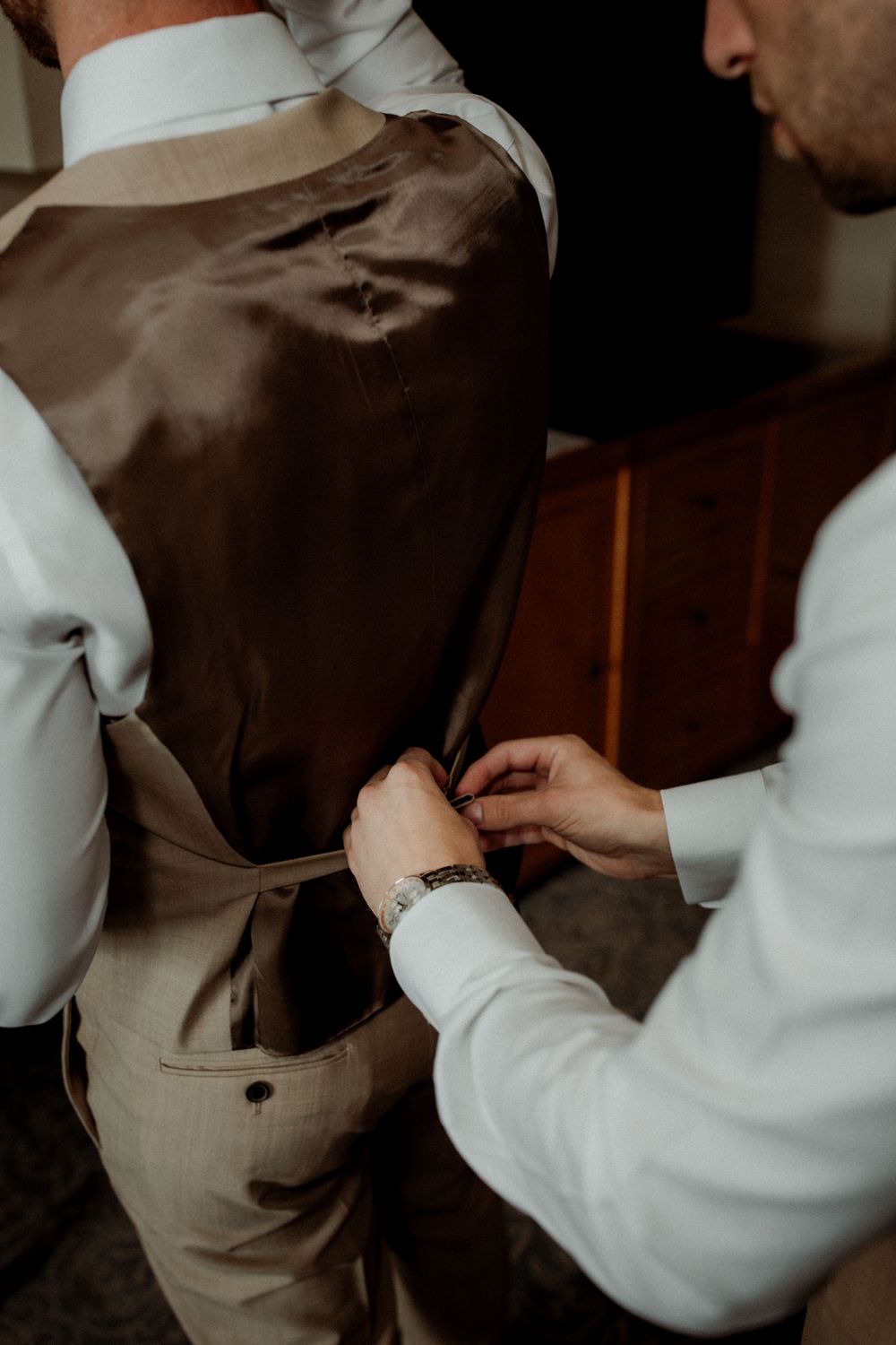 Someone helping adjust the back straps of a tan vest during formal wear preparation.
