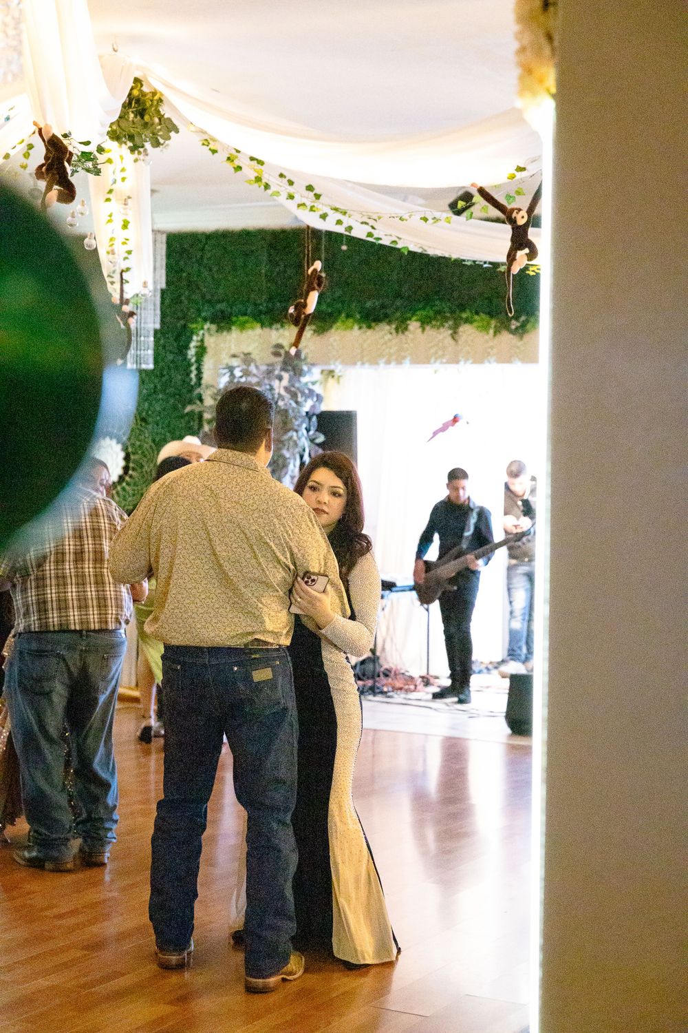 Couples slow dancing at an indoor event decorated with white drapes and greenery while a band performs in the background.