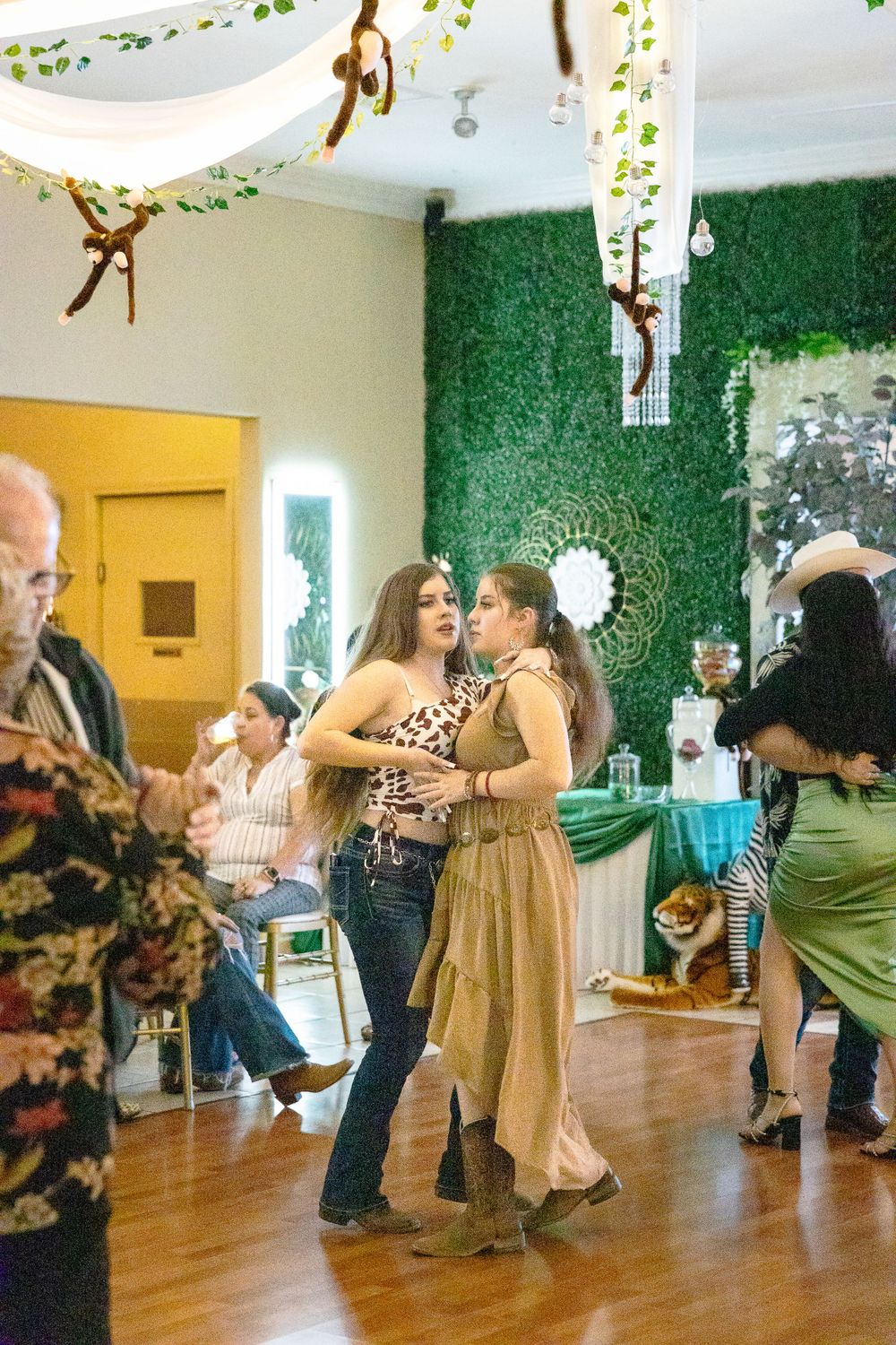 Party guests dancing in a venue decorated with hanging greenery and warm lighting.