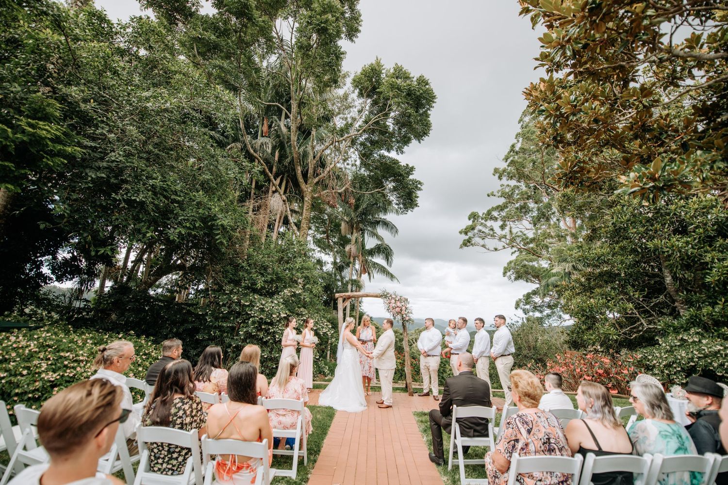 Wedding ceremony guests celebrate under lush green trees at an outdoor venue with white chairs.