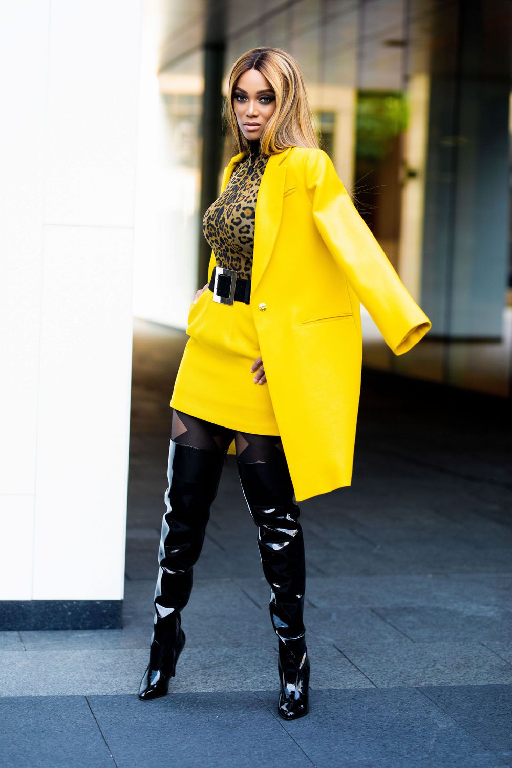 A woman wearing a yellow coat and skirt, leopard-print top, and black over-the-knee boots stands in an urban setting