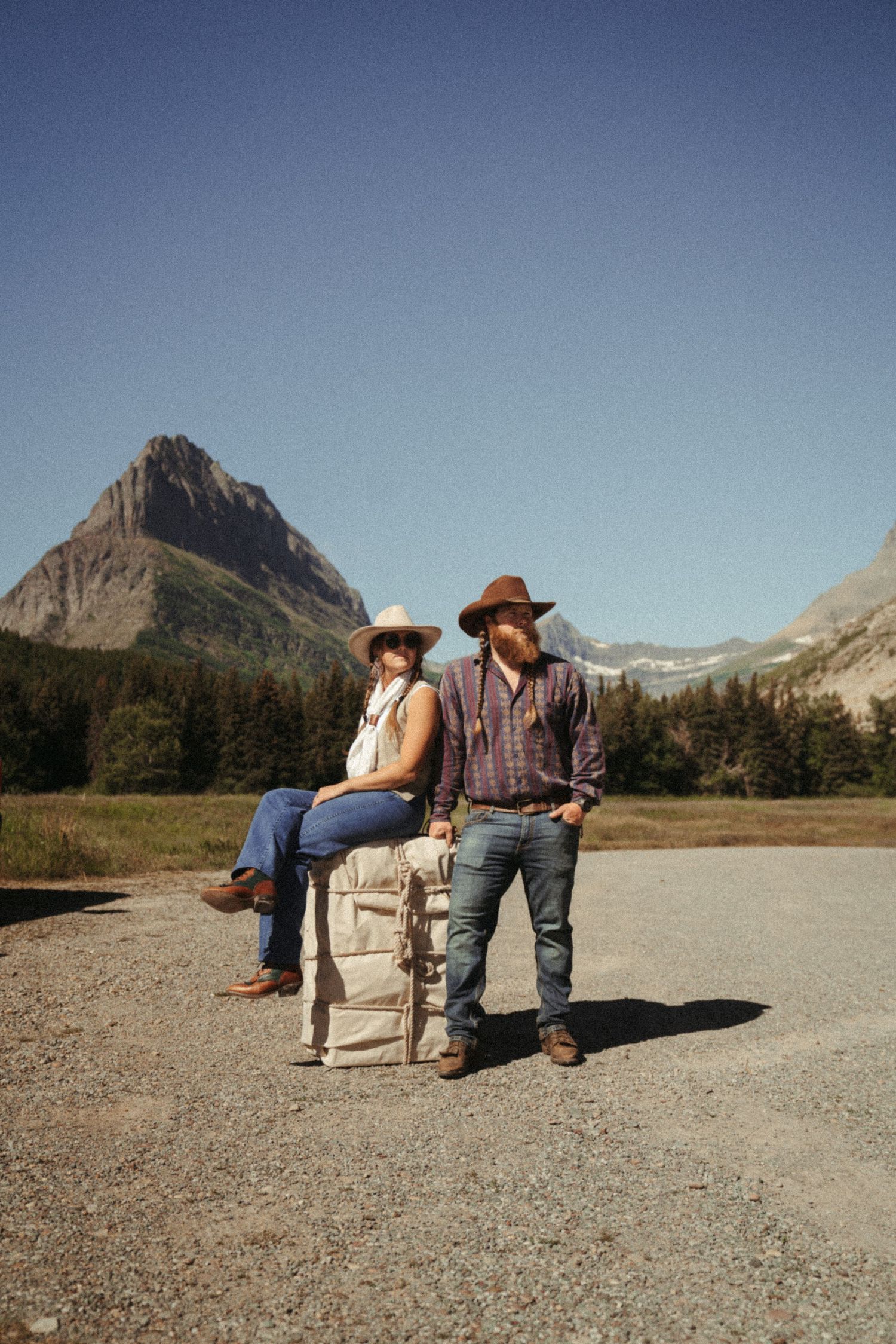 Two people in cowboy hats sit on mantied mule packer load in front of scenic mountain peaks in Glacier National Park.