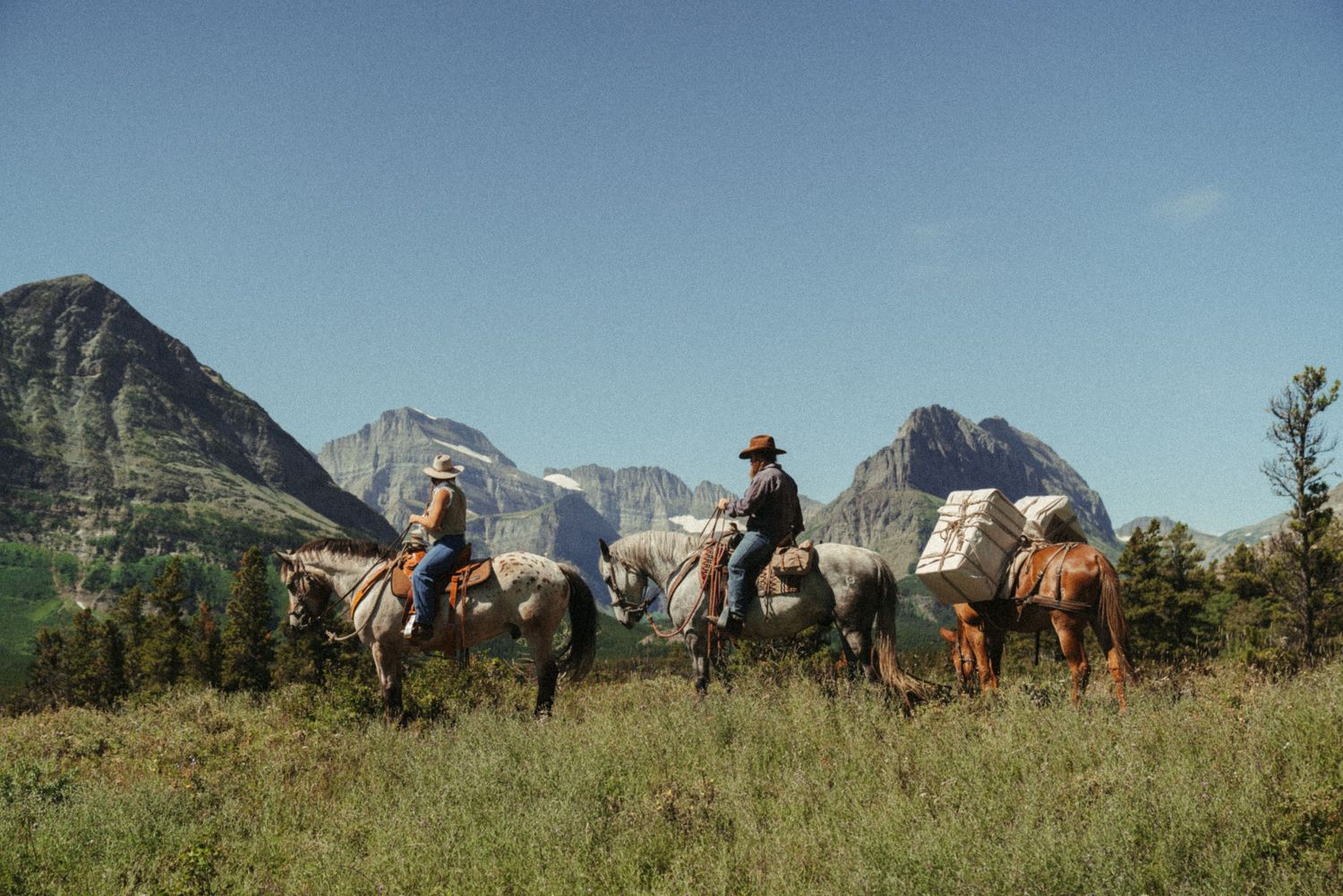 Horseback riders with pack mule travel along a scenic mountain trail with horses and gear against a backdrop of rugged peaks in Many Glacier